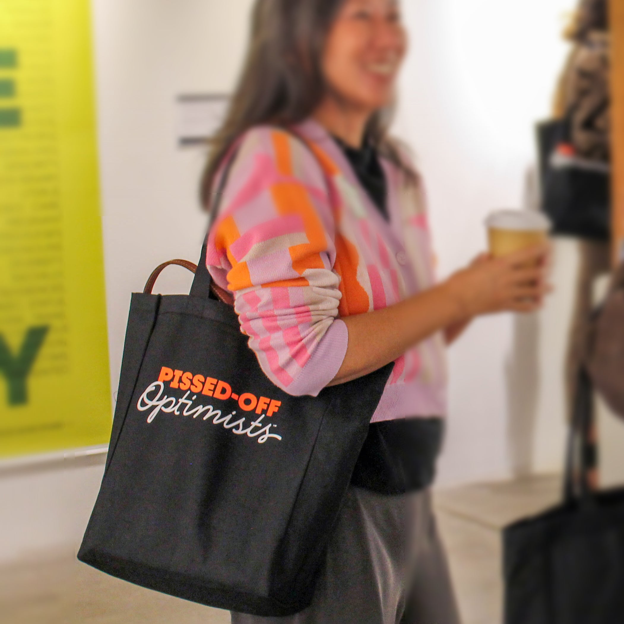 Person holding a black tote bag with 'PISSED-OFF Optimists' text, wearing a colorful cardigan.